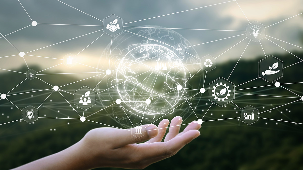 Hand holding digital globe with interconnected icons, including gears, plants, and graphs, symbolizing technology and sustainability against a blurred natural background.