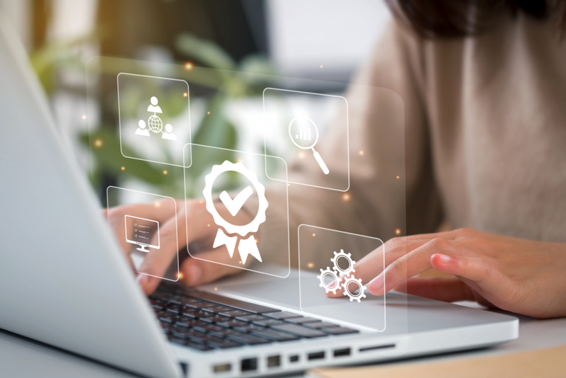 Person typing on a laptop with floating icons: globe, magnifying glass, checkmark ribbon, computer monitor, and gears. Concept of digital technology and workflow.