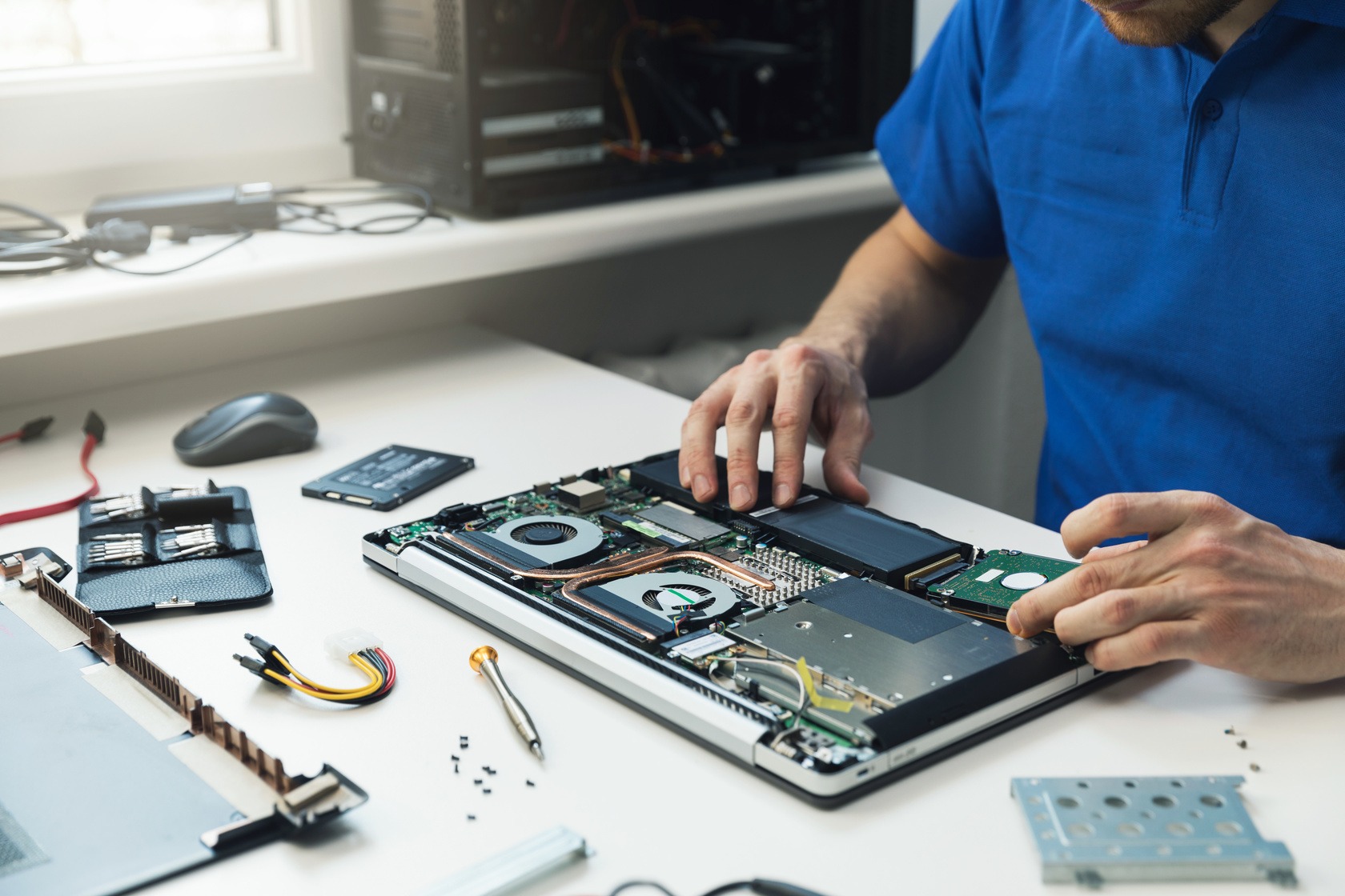 A person in a blue shirt meticulously repairs a laptop, revealing internal components like the fan, battery, and hard drive. Nearby, tools and screws are neatly organized on the desk—a real one-stop shop for all your tech repair needs.