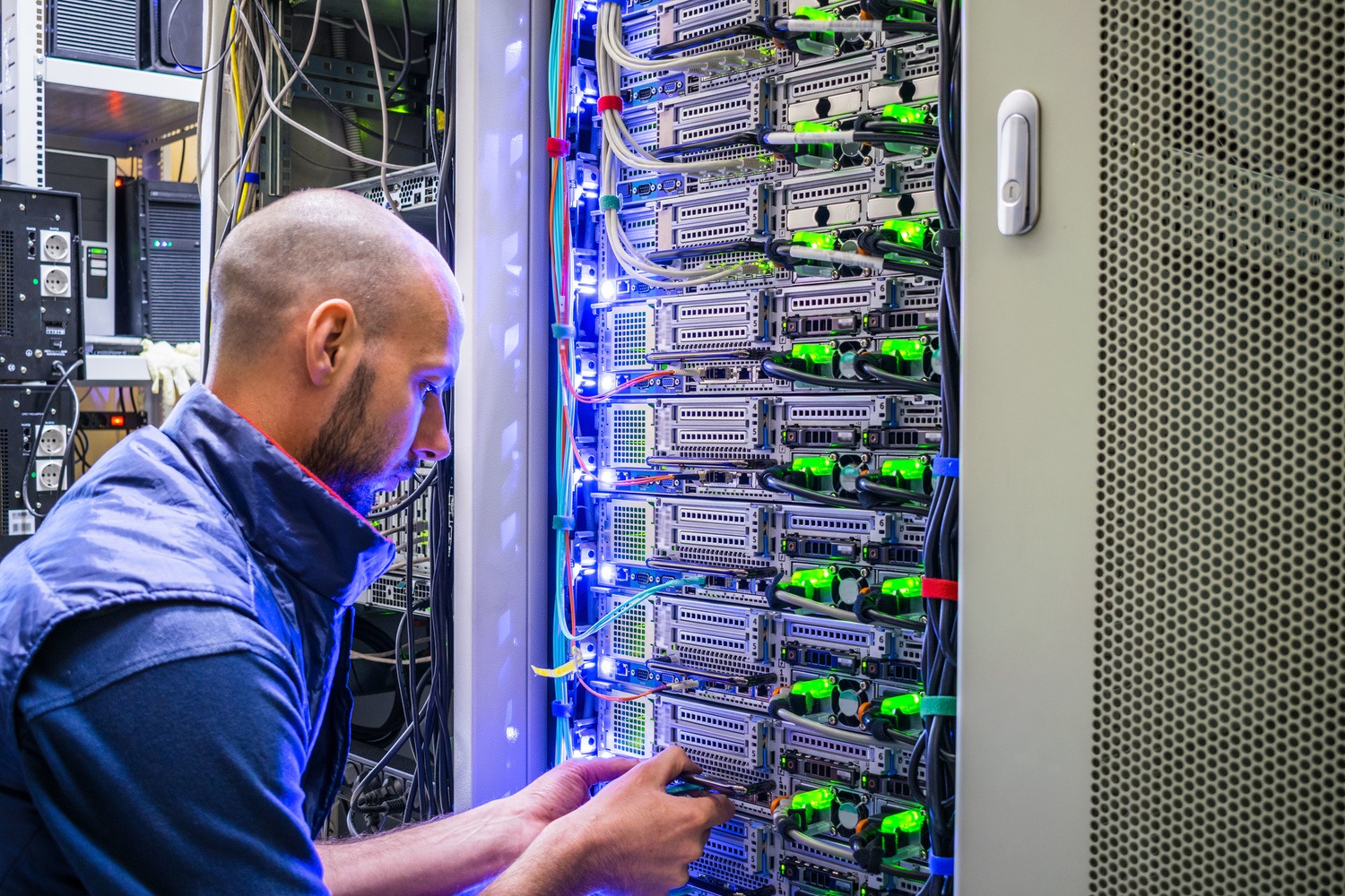 A person is meticulously working on a server rack, organizing various cables amidst the blinking lights in a data center, ensuring everything adheres to NCS standards.