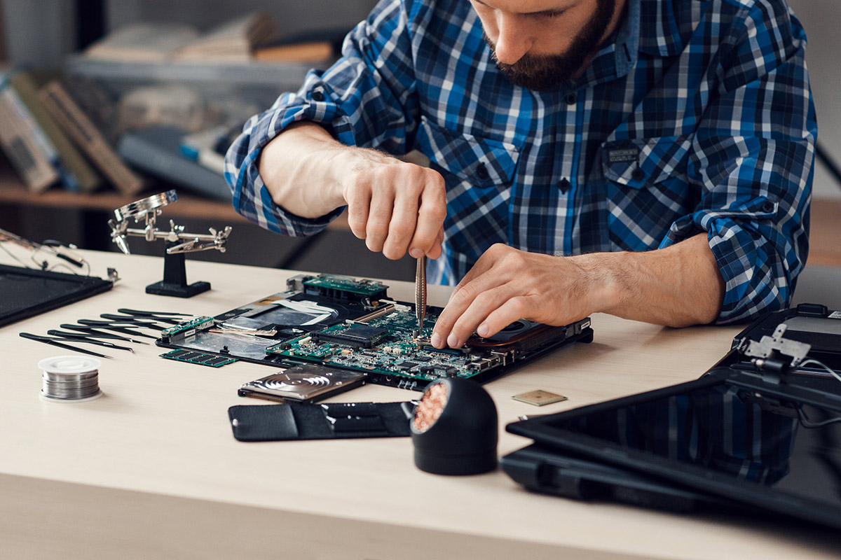 Person in a plaid shirt working on a disassembled laptop at a desk with tools and electronic components.