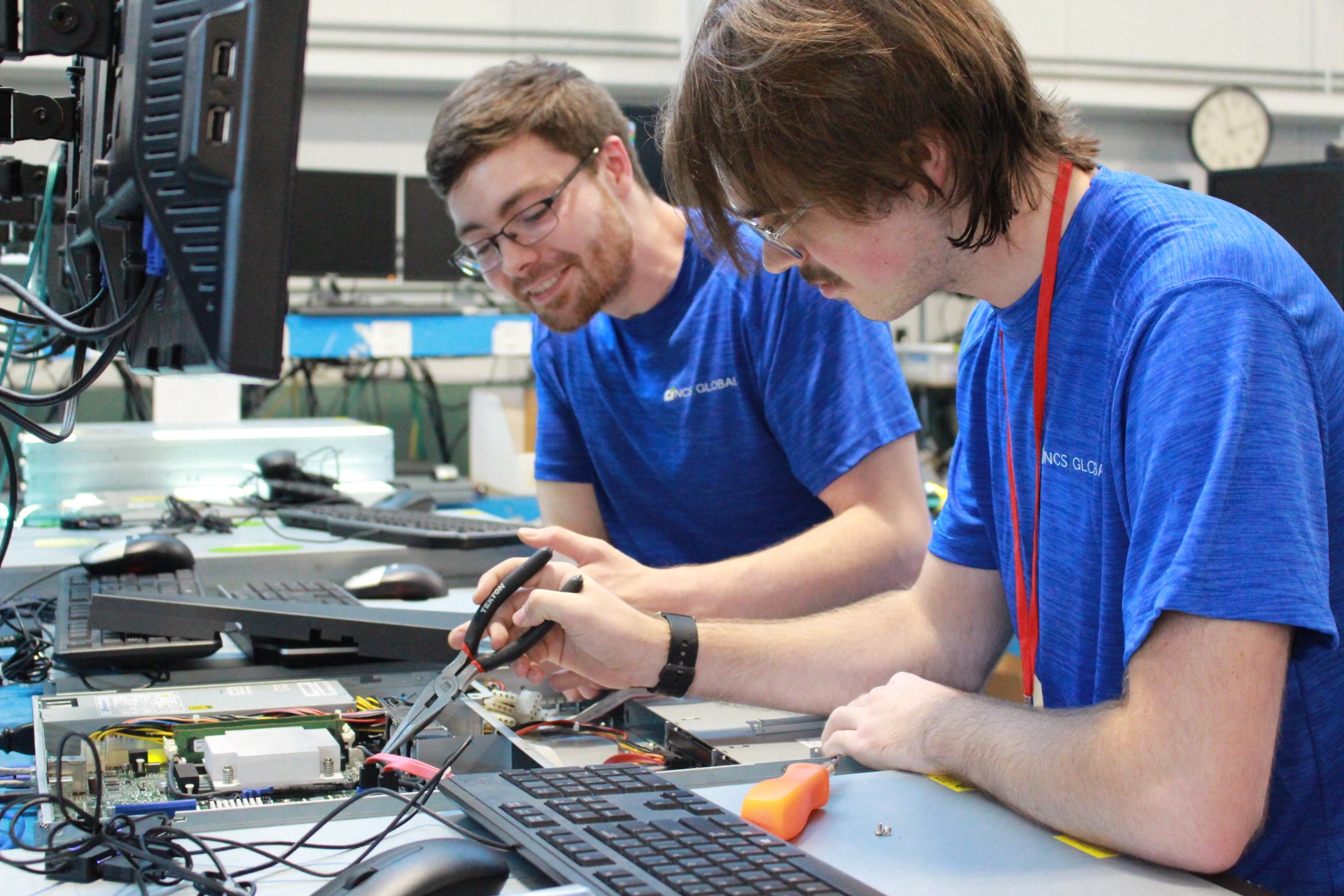 Two technicians in blue shirts, part of the NCS Global Internship Program, work on computer hardware at a desk surrounded by monitors and keyboards. One uses pliers while the other observes attentively.