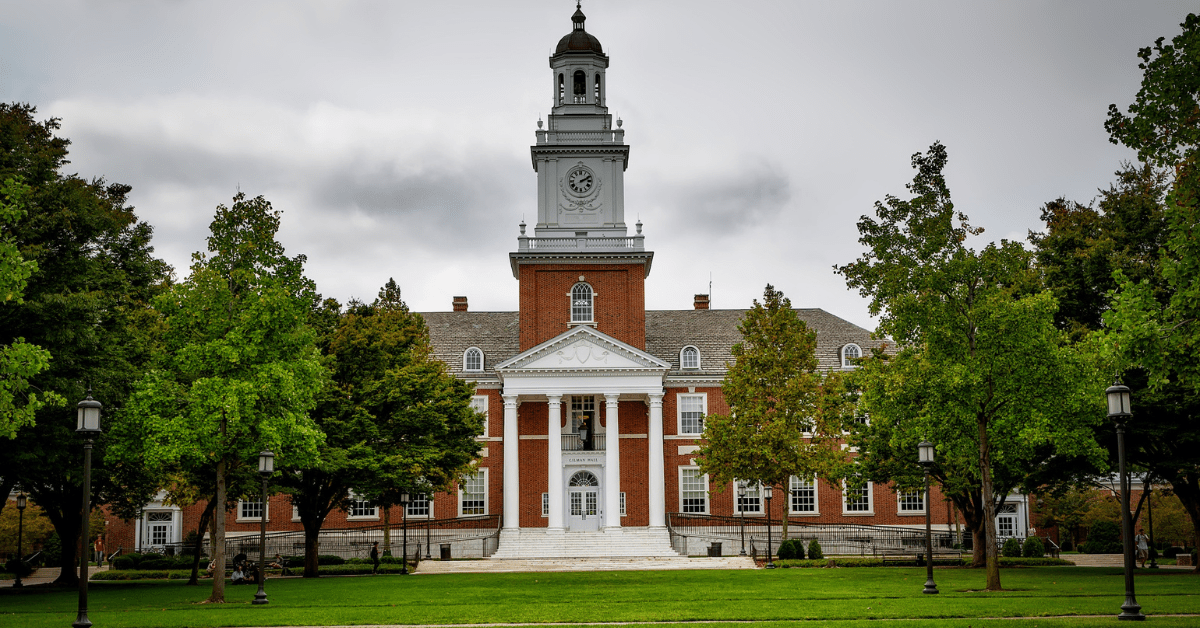 A large brick building with a central white clock tower and columns stands among lush trees, embodying the legacy of esteemed academic organizations. Under a cloudy sky, it serves as a beacon of knowledge, where client success stories unfold and value returns are maximized.