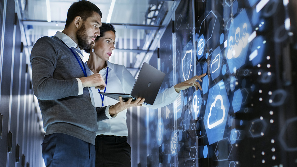 Two people in a server room examine a digital interface on a glass panel. One holds a laptop while the other points to cloud icons and data visuals displayed on the screen.