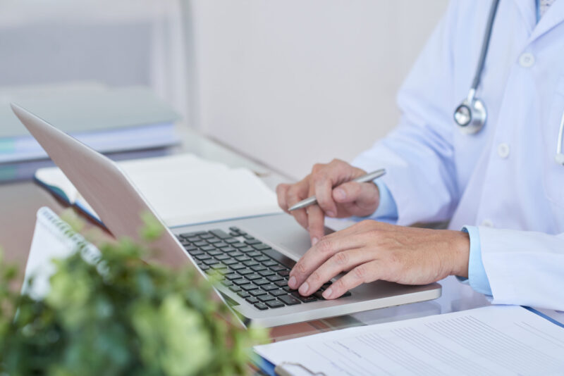 A doctor in a white coat uses a laptop at a desk cluttered with papers and charts, symbolizing the shift in healthcare as data centers streamline patient information. A stethoscope hangs around their neck, while a potted plant adds a touch of green to the bustling scene.