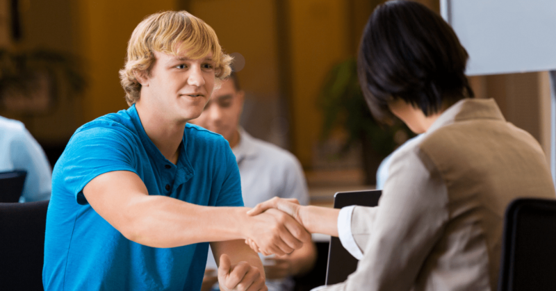 In an office setting, two people engage in a cordial handshake across the table during the NCS Global Career Fair. One person sports a blue shirt, while the other dons a beige jacket, highlighting their professional interaction against this dynamic backdrop.