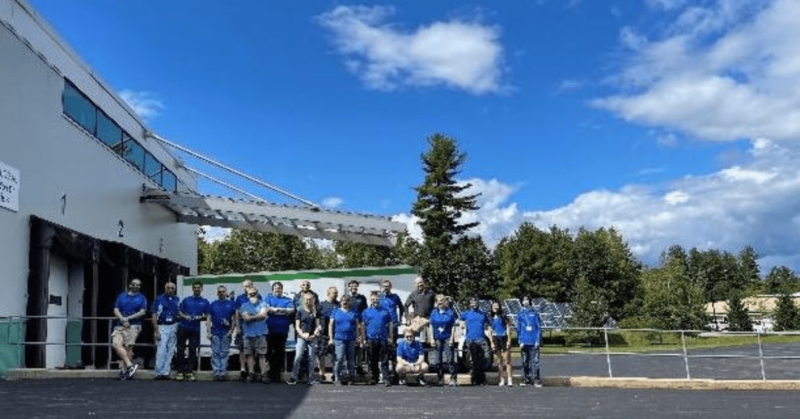 A group of people in blue shirts stands outside a building with a white overhang, celebrating NCS Global's 10th Anniversary. Trees create a serene backdrop under the partly cloudy sky as Martin Estes joins the gathering.