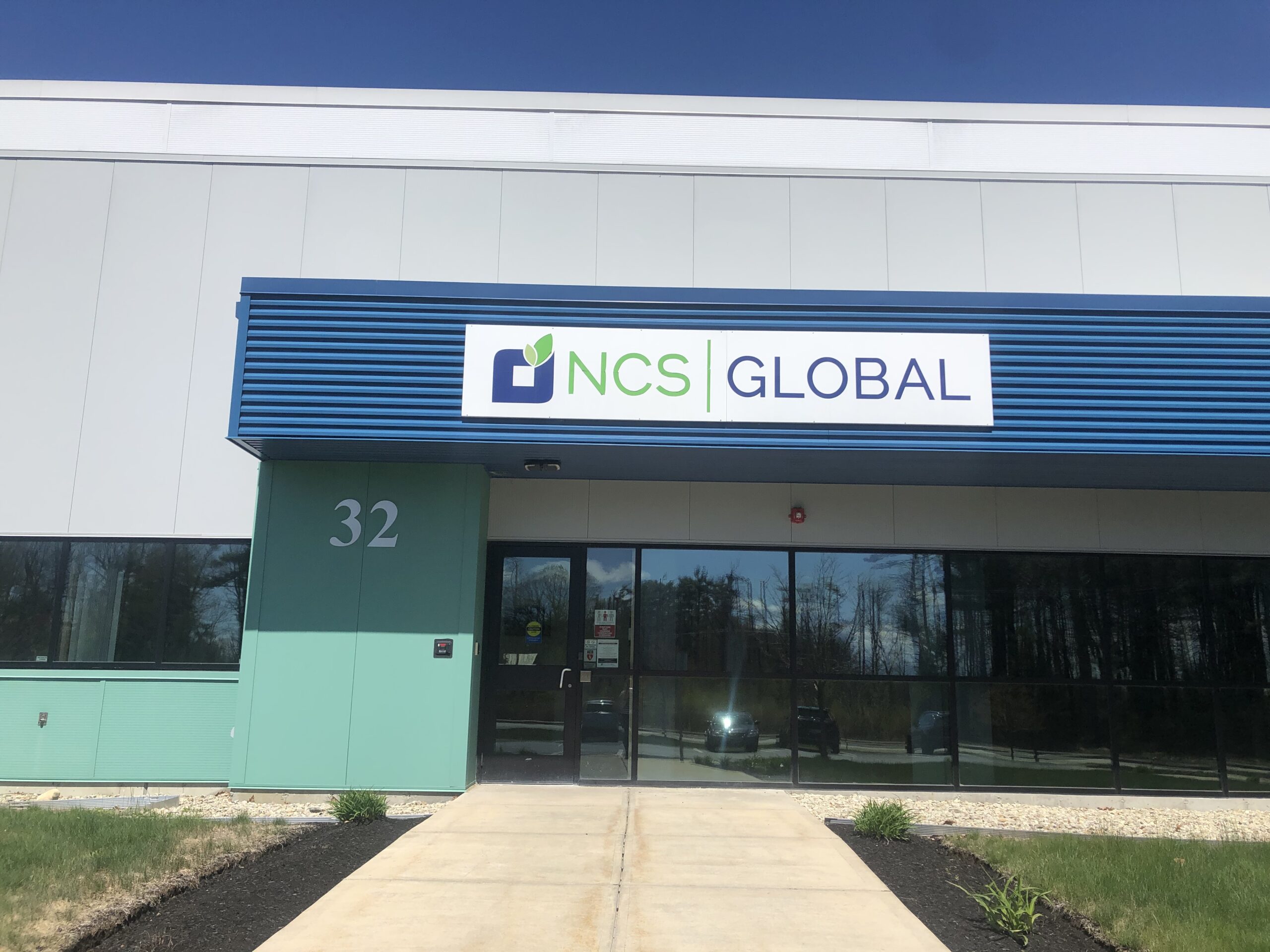 Exterior view of a building featuring a sign with the logo and text "NCS Global," proudly celebrating its 28th Anniversary. The building, marked with the number 32, has a walkway leading to the glass entrance door, highlighting years of success and commitment.