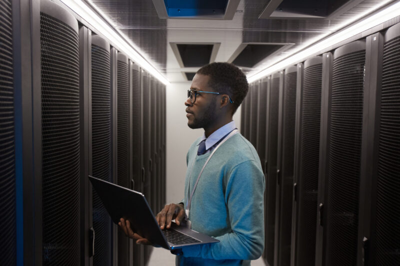 A person holding a laptop stands in a server room lined with black server racks, ensuring seamless customer service operations.