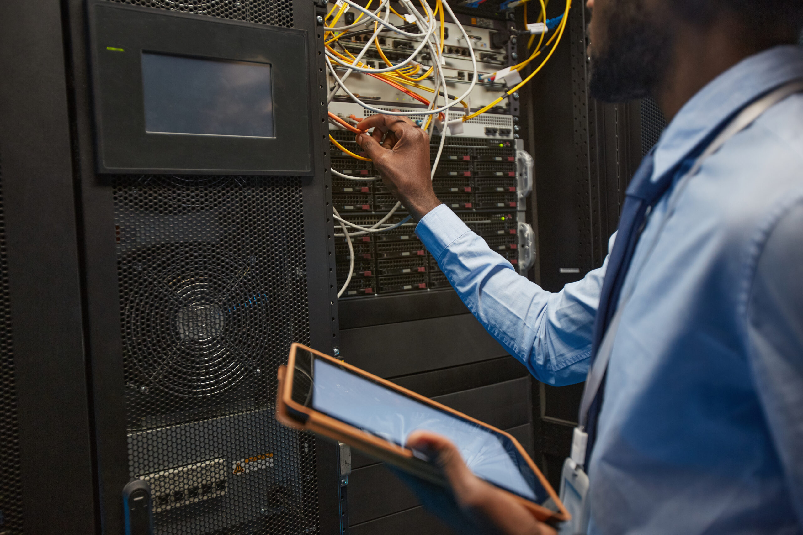 A person in a blue shirt is holding a tablet, implementing asset recovery solutions while checking cables in the server room to deliver results efficiently.