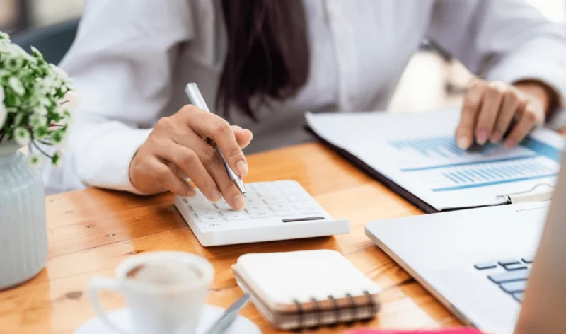 Person calculating numbers with a calculator, documents, and laptop on a wooden desk