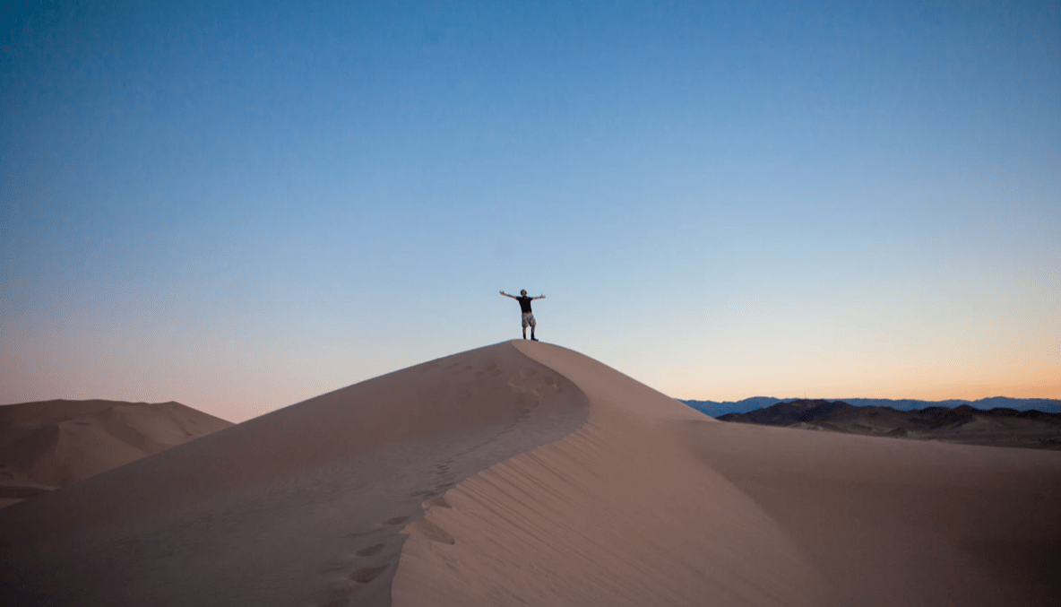 A person stands with arms outstretched atop a sand dune at sunset, embracing the serene beauty of the clear sky. This moment of reflection symbolizes continuous improvement, as they rise above challenges like the ever-shifting sands.