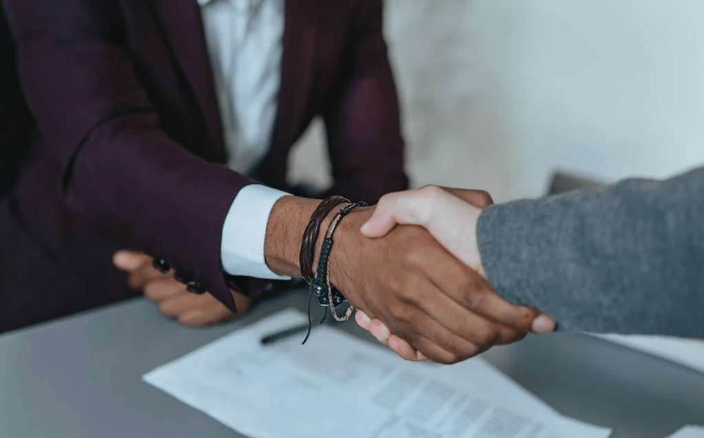 Two people shaking hands over a table with documents, signaling a successful agreement. One, adorned in a dark suit and bracelets, exudes confidence in selecting the right ITAD Vendor. The other person wears a gray suit, reflecting professionalism in their IT asset disposition discussion.