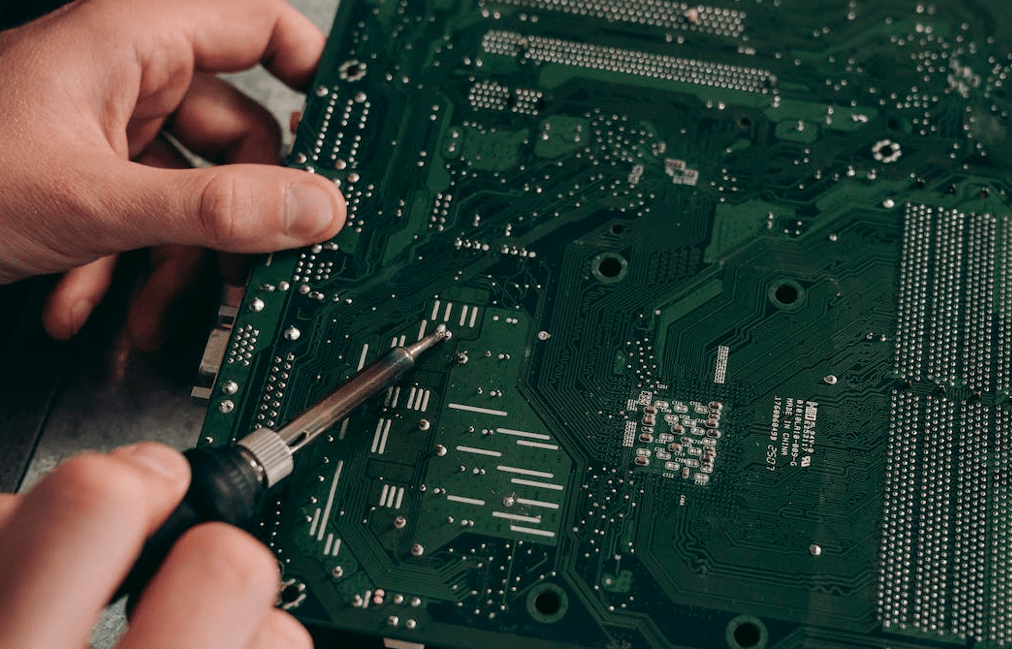 Hands meticulously soldering components on a green circuit board, with a soldering iron in focus, embody the spirit of improving North America's approach to technology and sustainability.