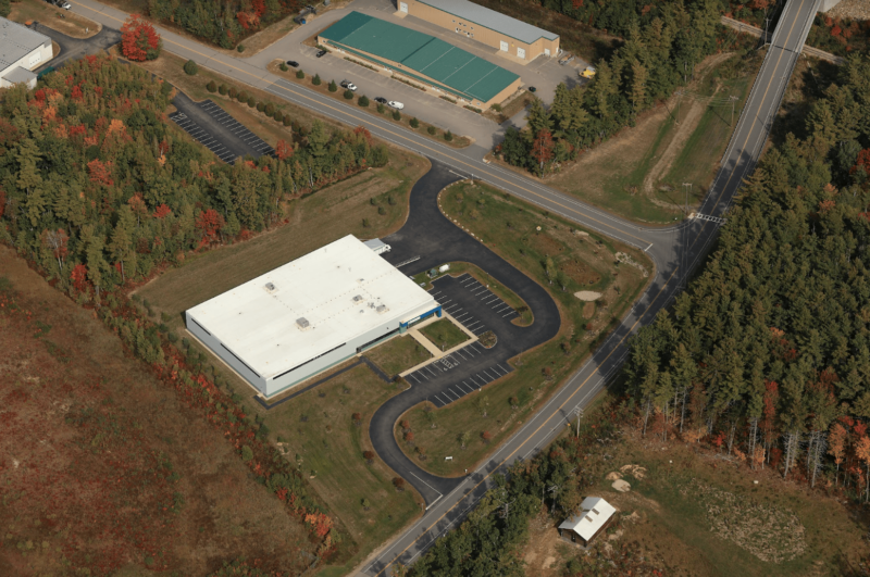 Aerial view of NCS Global's white-roofed industrial building with a winding driveway, surrounded by parking lots, trees, and roads at an intersection. Nearby is a green-roofed structure. An ideal setting for their end-of-year message backdrop.