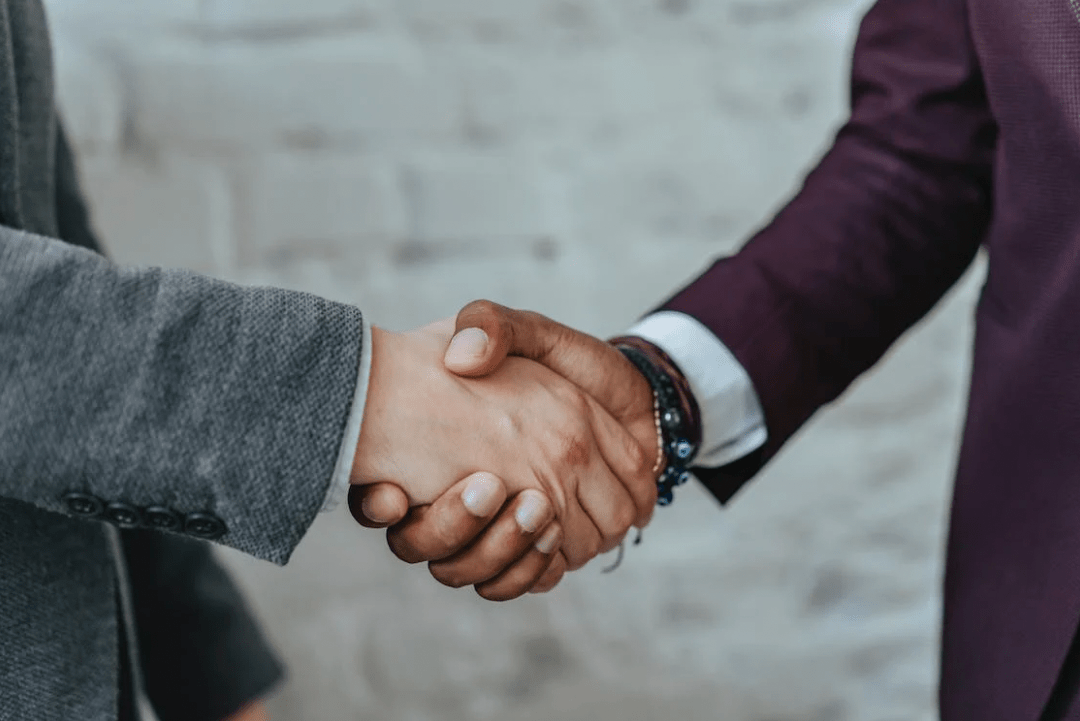 In front of a light background, two individuals in formal attire shake hands; one in a gray suit and the other in a dark purple suit, symbolizing a successful partnership between niche company service providers and big company performance experts.