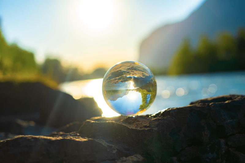 A glass globe on a rock reflects a sunny landscape with water, trees, and a mountain. The sun shines brightly in the background, casting warmth and gratitude like the perfect "thank you" to all who admire its beauty.