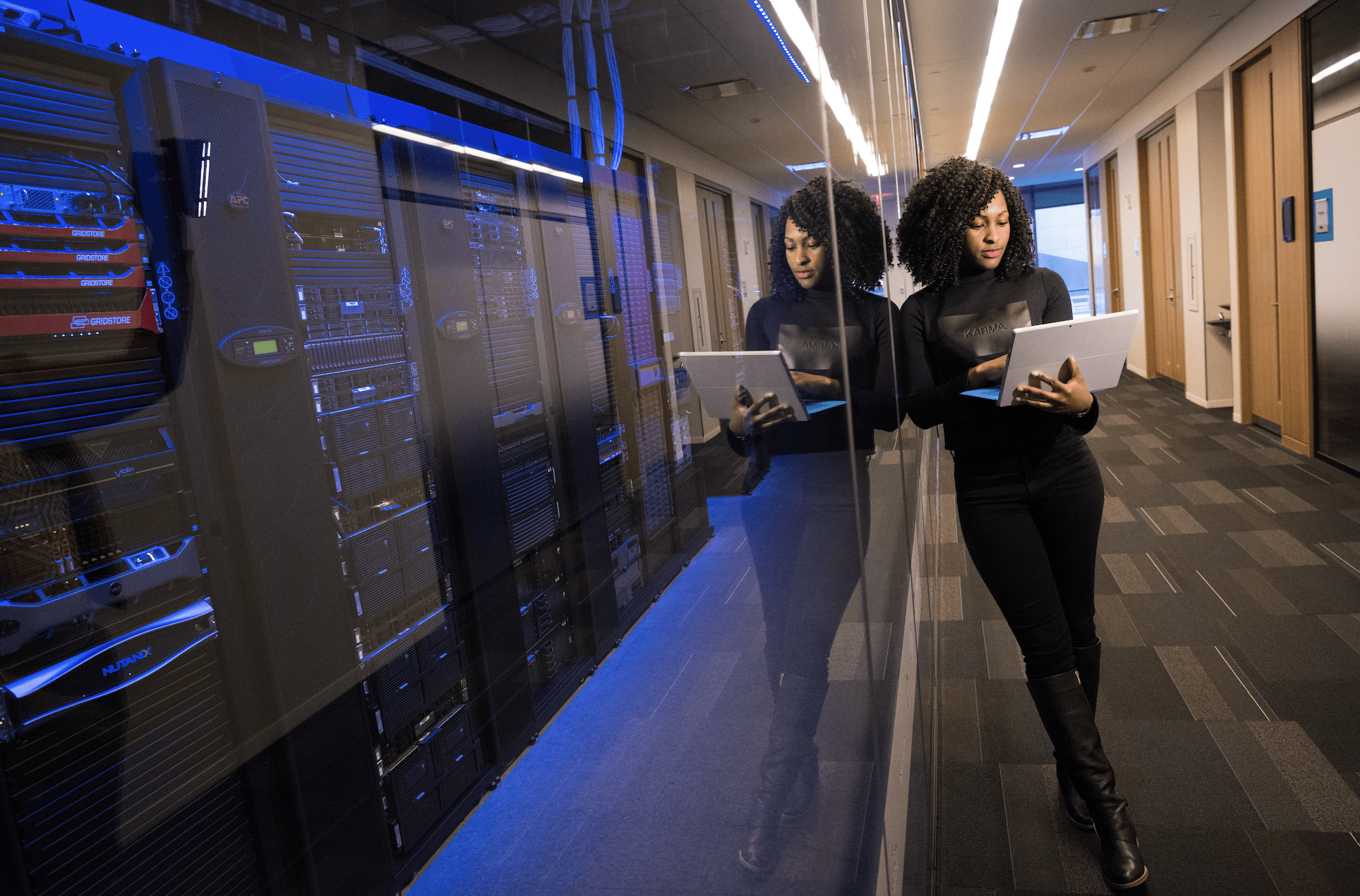 A woman stands with a laptop beside a glass wall, reflecting server racks in a data center hallway, where IT assets are monitored for efficient lifecycle management.