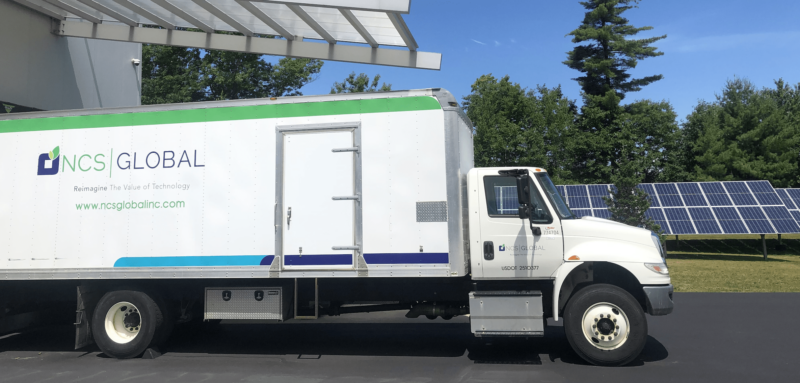 A white truck adorned with NCS Global branding proudly sits on a driveway beside a solar panel installation, trees swaying gently in the background—all under a clear blue sky. This picturesque scene also celebrates National Truck Driver Week, honoring those who keep America moving.