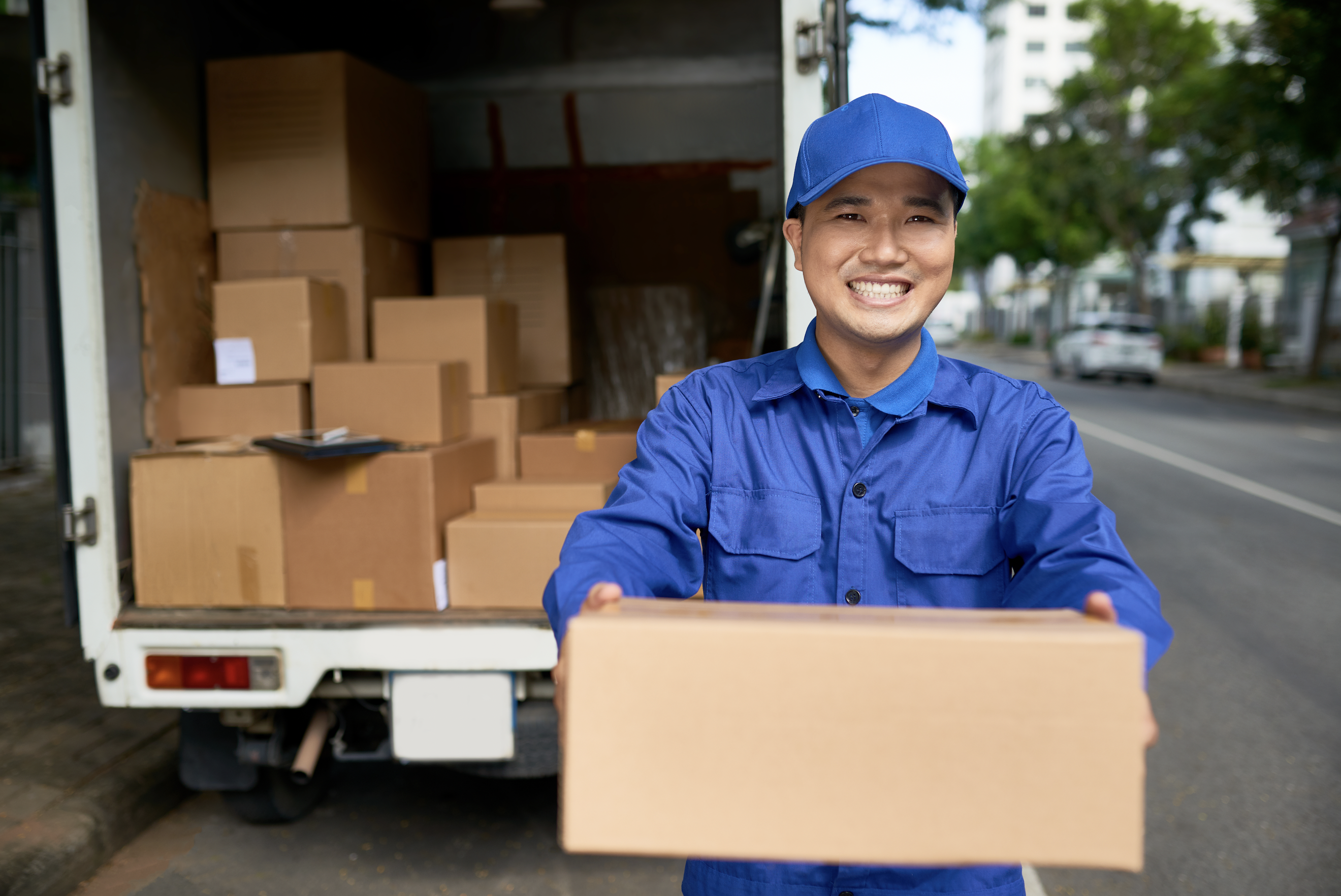 A delivery person in a blue uniform smiles, holding a cardboard box beside an open truck filled with more, showcasing the ins and outs of reverse logistics on a bustling city street.