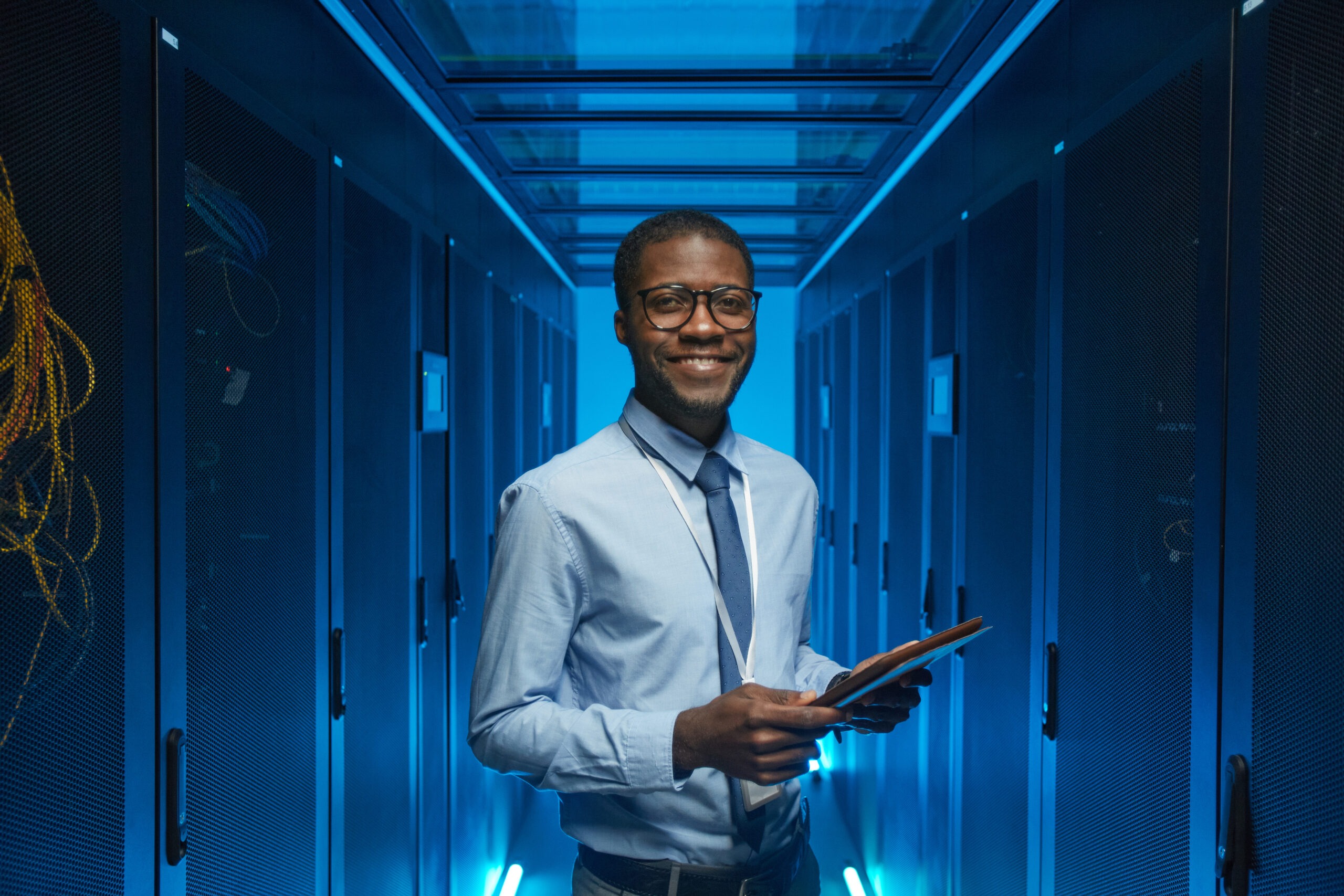 A man in a shirt and tie stands smiling in a server room, holding a tablet. Blue lighting highlights the rows of server racks on either side, showcasing his winning approach to ensuring impeccable data security.