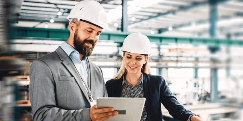 Two people in hard hats and business attire review industry trends on a tablet in an industrial setting.