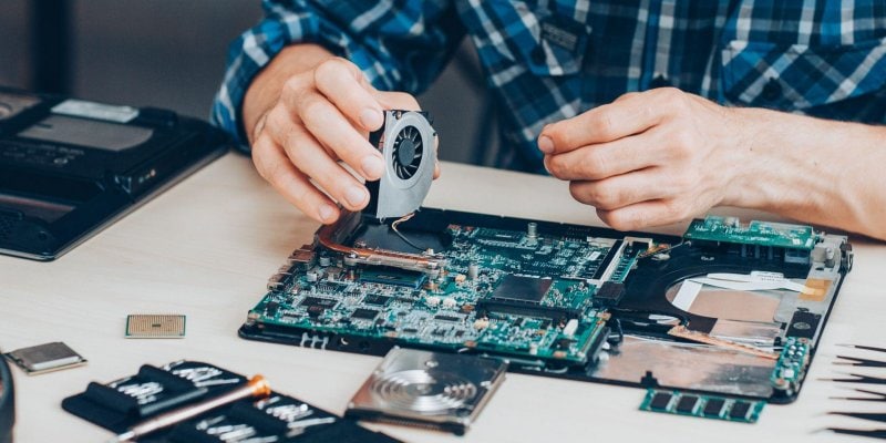 man placing a fan on a motherboard A person is assembling or repairing a laptop motherboard, holding a cooling fan. Various electronic components and tools are spread out on the table—perfect for those considering outsource IT asset repairs for efficient device maintenance.