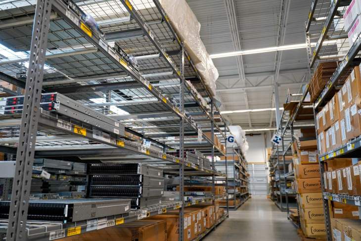 In a warehouse aisle brimming with the latest trends in ITAD industry advancements, metal shelves hold cardboard boxes and various equipment, all illuminated by bright overhead lighting, offering a glimpse into the future of logistics in 2025 from a low angle.