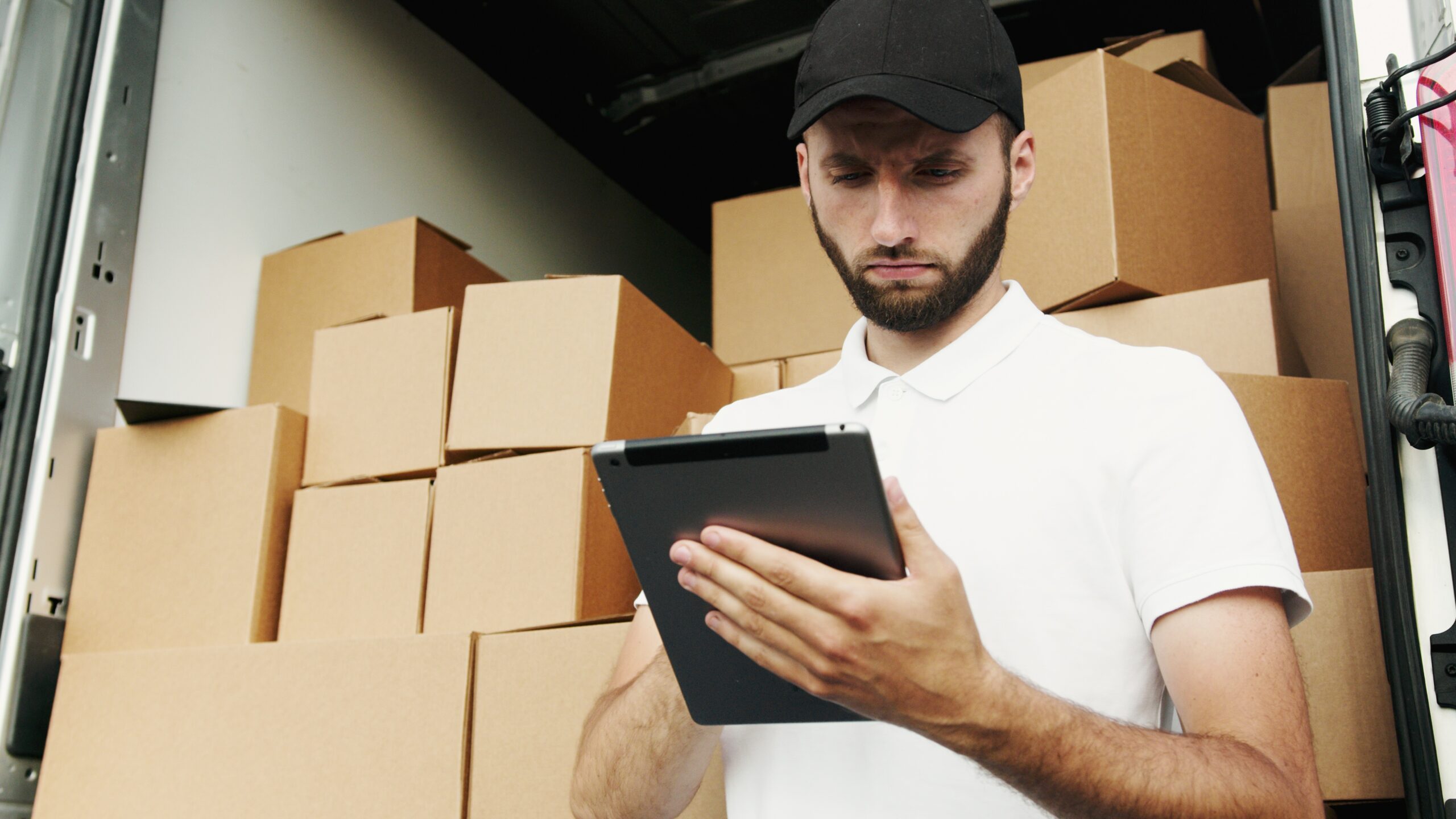 A man from a global shipping company, donning a cap and white polo shirt, stands before a stack of cardboard boxes, intently examining a tablet.