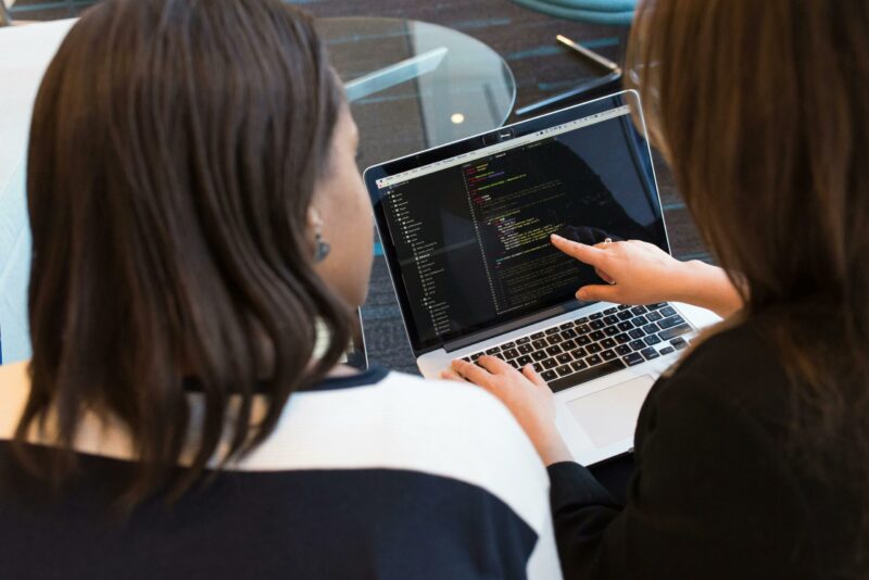 Two people sit at a table, looking at a laptop screen displaying lines of code, with one person pointing to it as they discuss the right type of data destruction strategies for their company.