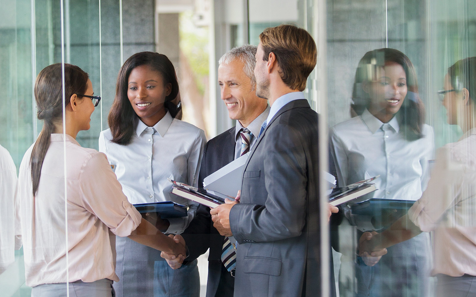 Four business professionals in formal attire are gathered in a hallway. Two are shaking hands while holding folders. The other two are observing and smiling.